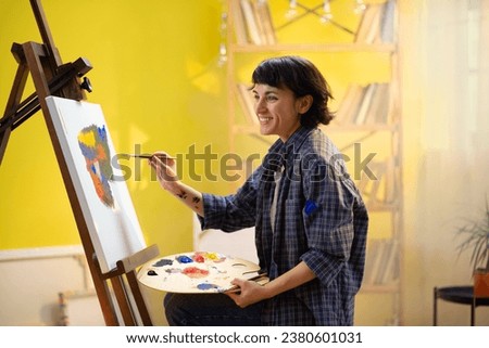 A beautiful artistic woman with a very beautifully structured face is painting something gracefully with her hand and paintbrush while looking very happy