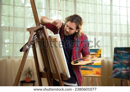 A very artistic looking man with medium hair and a beard is looking quite pleased with himself as he paints something onto a big canvas