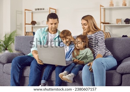 Happy young family with two kids girl and boy sitting on sofa using modern laptop together. Smiling parents resting on couch enjoying weekend watching funny cartoon online or talking on video call.