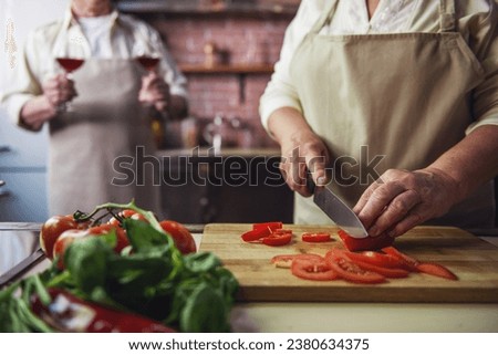 Cropped image of beautiful senior couple in aprons drinking wine and chopping tomato while cooking together in kitchen
