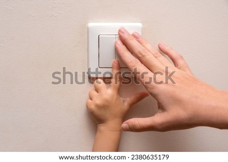 Mom teaches her kid to turn on the light inside the house or room. Parental control. Woman's and kid hands on a light switch Royalty-Free Stock Photo #2380635179