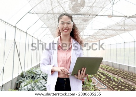 Happy smiling beautiful Asian botanist scientist woman in lab coat holding laptop computer, female biological researcher working on experimental plant plots in greenhouse. Biology Agricultural Science Royalty-Free Stock Photo #2380635255