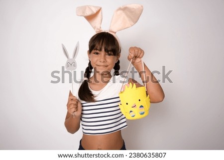 Photo of joyful brunette girl in toy rabbit ears smiling and holding colorful eggs isolated over white background