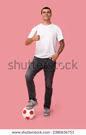 Handsome young man with soccer ball showing thumb-up gesture on pink background