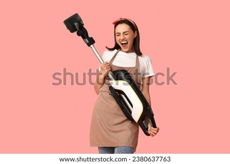 Portrait of happy young housewife in apron with vacuum cleaner on pink background Royalty-Free Stock Photo #2380637763