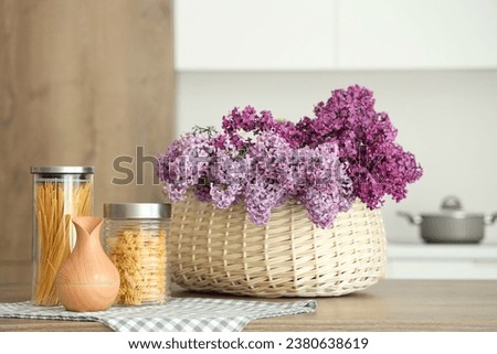 Basket with lilac flowers and jars of raw pasta on table in light kitchen Royalty-Free Stock Photo #2380638619