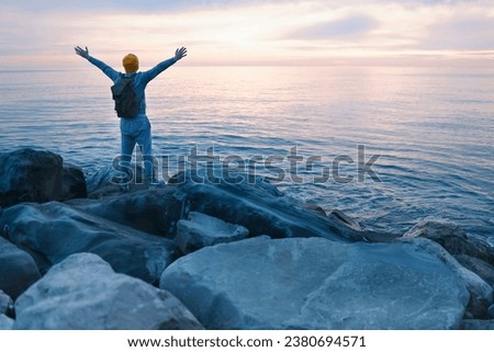 Young man in casual clothes sits on the rocks near the sea. Caucasian guy in a blue sweater and hat holds his backpack and looking at the calm sea. View from the back. The concept of nature and peace. Royalty-Free Stock Photo #2380694571