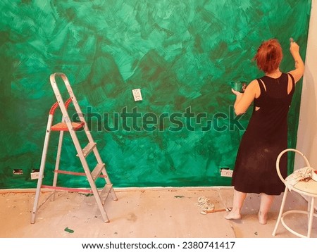 Red-haired woman takes advantage of her free time to paint her bedroom wall green, exercising her artistic talent to create textures on the wall.