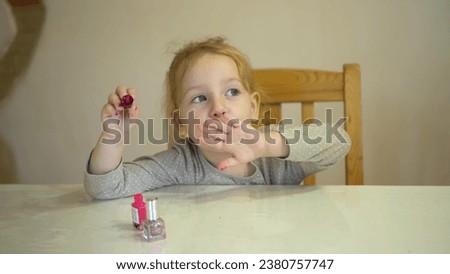 A little girl paints her nails with pink polish. The girl, having put on her makeup, blows on her nails to let them dry. Royalty-Free Stock Photo #2380757747