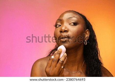 African American woman holding makeup egg sponge, powdering, making modern daily make-up, isolated on colorful background. Skin care and beauty concept Royalty-Free Stock Photo #2380764693