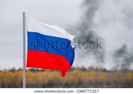The white-blue-red flag of the Russian Federation is flying in the wind against the background of black smoke in a cloudy sky.