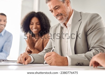 Happy old businessman executive investor banker signing legal business document agreement contract at board meeting with attorneys lawyers advisers diverse law team sitting at office table. Close up