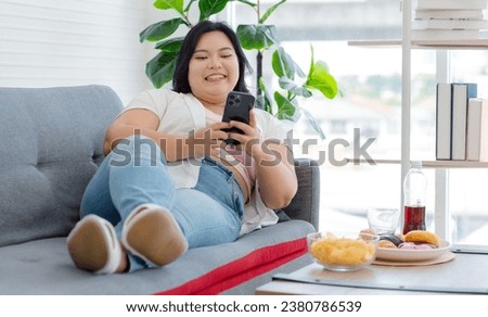 Asian young cheerful overweight oversized fat chubby plump unhealthy female teenager in casual outfit laying lying down smiling on cozy sofa holding croissant taking selfie photo via smartphone.