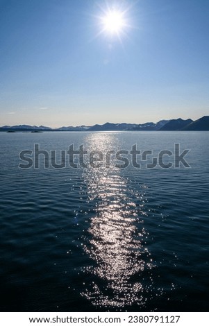 Sun starburst on a blue nature background of calm ocean water and blue sky separated by an eroded rocky mountain range of Gnalodden, arctic expedition tourism around Svalbard
