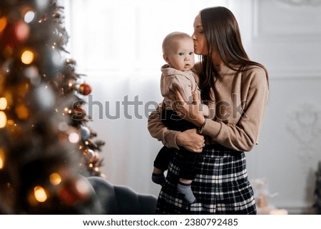A happy young mother holds her little son in her arms against the backdrop of a beautifully decorated New Year tree and kisses him. Christmas family atmosphere. Beautiful portrait of mother and son