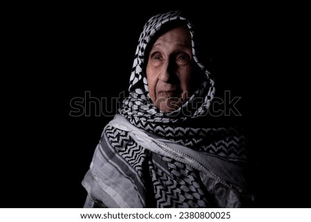 portrait of an old lady in dark background wearing white palestinian keffiyeh with smile on her face looking for freedom and hope to Right of Return for Palestinians Royalty-Free Stock Photo #2380800025