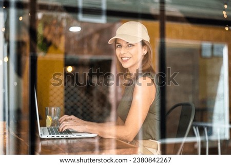 Freelance smiling woman working in a coffee shop with a laptop on her day off. Woman in coffee shop using technology. Photographed through the window. lifestyle