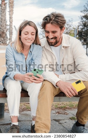 Vertical portrait of a young adult friends using their cellphones, browsing on internet enjoying free time sitting on bench outside. Happy caucasian couple having fun together with their smart phones