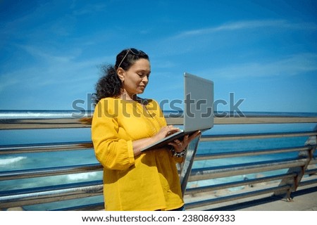Purposeful female developer, trader, manager online working on laptop outdoor. Beautiful business woman typing text on computer keyboard, planning startup, standing against a seascape background