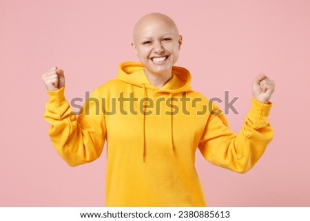 Young bald smiling caucasian happy positive woman 20s without hair wearing yellow casual sweatshirt shirt do winner gesture clench fist celebrating isolated on pastel pink background studio portrait