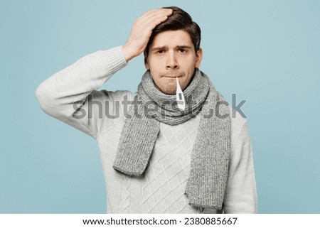 Young sad ill sick man wear gray sweater scarf hold keep thermometer in mouth isolated on plain blue background studio portrait. Healthy lifestyle disease virus treatment cold season recovery concept