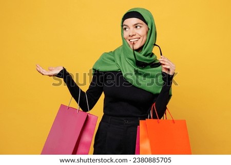 Young smiling happy muslim woman wear green hijab abaya clothes hold shopping package bags point aside on area isolated on plain yellow background studio. Black Friday sale buy day, uae islam concept