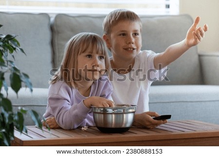 Cute joyful brother and sister are sitting at the table with a bowl of popcorn and watching TV. The concept of children's leisure, pastime.