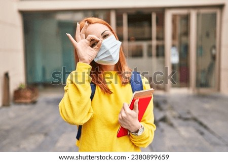 Young woman wearing safety mask and student backpack holding books smiling happy doing ok sign with hand on eye looking through fingers 