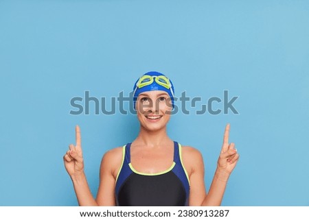 Young happy swimmer girl in swimming costume with goggles on head stands on blue background with fingers pointed up, professional sport concept, copy space. 