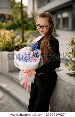 Young darkhair girl in black outfit holding huge bouquet of different fresh flowers in wrapping paper on blurred background. Mix of delicate flowers in woman's hands. Outdoors Royalty-Free Stock Photo #2380919427
