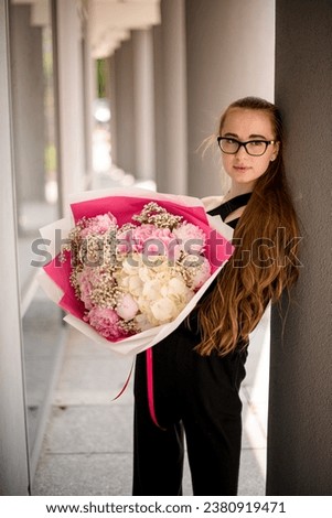 Portrait of stylish young girl wearing blak clothes holding bouquet of flowers, posing in between columns of modern building. Fresh mix of peonies, hydrangea and gypsophila flowers in woman's hands