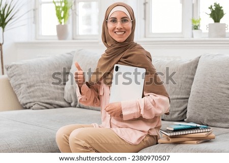 Satisfied and happy Muslim woman in hijab and glasses siting on sofa in cozy living room, holding tablet ,shows thumbs up. Female student, tutor or teacher is ready to start online learning remotely. 