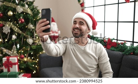 Young hispanic man make selfie by smartphone sitting on sofa by christmas tree at home