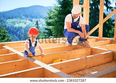 Father with toddler son constructing wooden frame house. Man instructing his son on measurement of distance using tape measure on construction site, wearing helmets and blue overalls on sunny day. Royalty-Free Stock Photo #2381027287