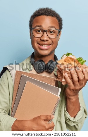 Vertical image of cheerful handsome guy with curly hair holds tasty sandwich and notebooks smiles gladfully dressed in casual clothing wears stereo headphones around neck isolated over blue background Royalty-Free Stock Photo #2381052211