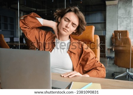 Image of woman with tired face, sits with laptop in office, feels tension in neck, pain in muscles after working all day at workplace. Royalty-Free Stock Photo #2381090831