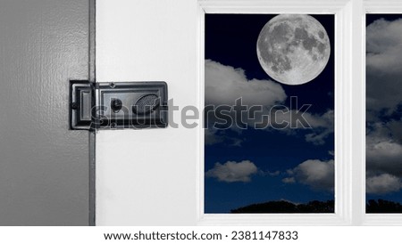 Close-up color photo of an old metal door lock on a white wooden door with window panels. A beautiful full moon and white fluffy clouds can be seen through the window. Copy space.