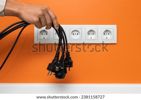 Human hand holding black cables in front of a five-way wall power socket on the orange wall. Electrical plugs gently hang from the palm Royalty-Free Stock Photo #2381158727
