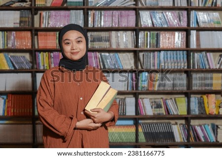 Portrait of Asian hijab woman holding book in front of library bookshelf. Muslim girl reading a book. Concept of literacy and knowledge