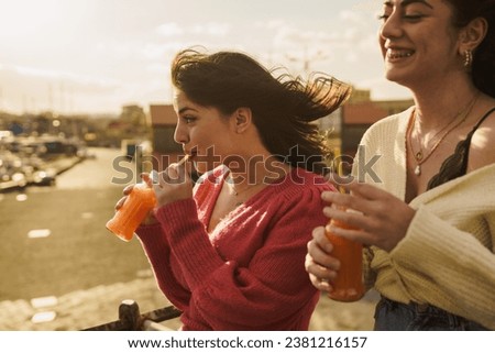 Two friends share a delightful moment at the harbor, wind tousling their hair. Their genuine smiles and vibrant drinks capture the essence of a perfect summer day spent in good company. Royalty-Free Stock Photo #2381216157
