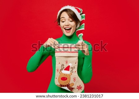 Young surprised fun shocked woman wear green turtleneck Santa hat posing hold look at stockings with gifts isolated on plain red background. Happy New Year 2024 celebration Christmas holiday concept