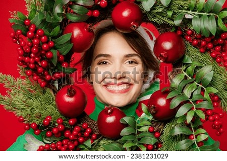 Close up young fun woman wear green turtleneck Santa hat posing hold in hand look through decorative wreath isolated on plain red background. Happy New Year 2024 celebration Christmas holiday concept