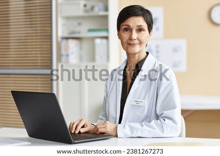 Portrait of smiling mature woman as female doctor using computer in clinic and looking at camera Royalty-Free Stock Photo #2381267273