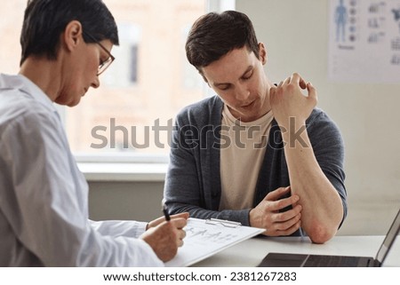 Portrait of young man showing skin rash to doctor during consultation in dermatology clinic Royalty-Free Stock Photo #2381267283