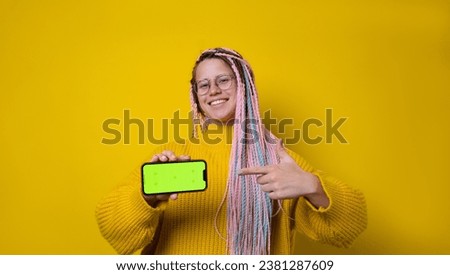 Excited young woman with afro braids pointing finger to blank phone green screen with copy space for app, isolated on yellow background. High quality photo