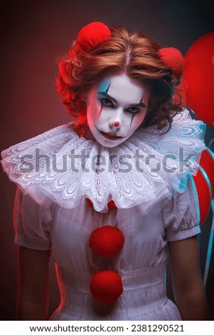 Scary Halloween. A clown girl in a white circus dress holds red balloons in her hand and looks gloomily at the camera. Dark studio background with red light.