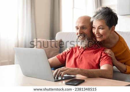 Portrait Of Happy Senior Couple Using Laptop At Home, Smiling Elderly Spouses Resting With Computer In Living Room, Shopping Online Or Watching Photos Together, Wife Embracing Husband From Behind