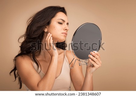 Stressed sad brunette young woman wearing beige top looking at mirror and touching her face over beige studio background, having skin problems. Acne, pimples, wrinkles, dull skin Royalty-Free Stock Photo #2381348607