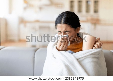 Cold And Flu Symptoms . Sick young indian woman covered in blanket blowing her running nose in a tissue while sitting on couch in living room, ill lady drinking hot tea, feeling unwell at home Royalty-Free Stock Photo #2381348975