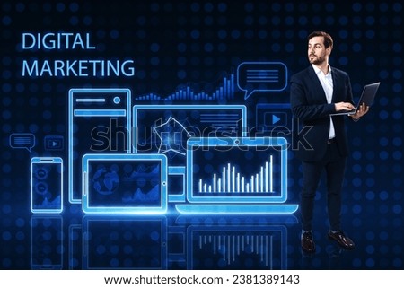 Attractive young european man using laptop while standing on blue pixel background with gadgets and business chart hologram. Digital marketing, finance, social network and online service concept